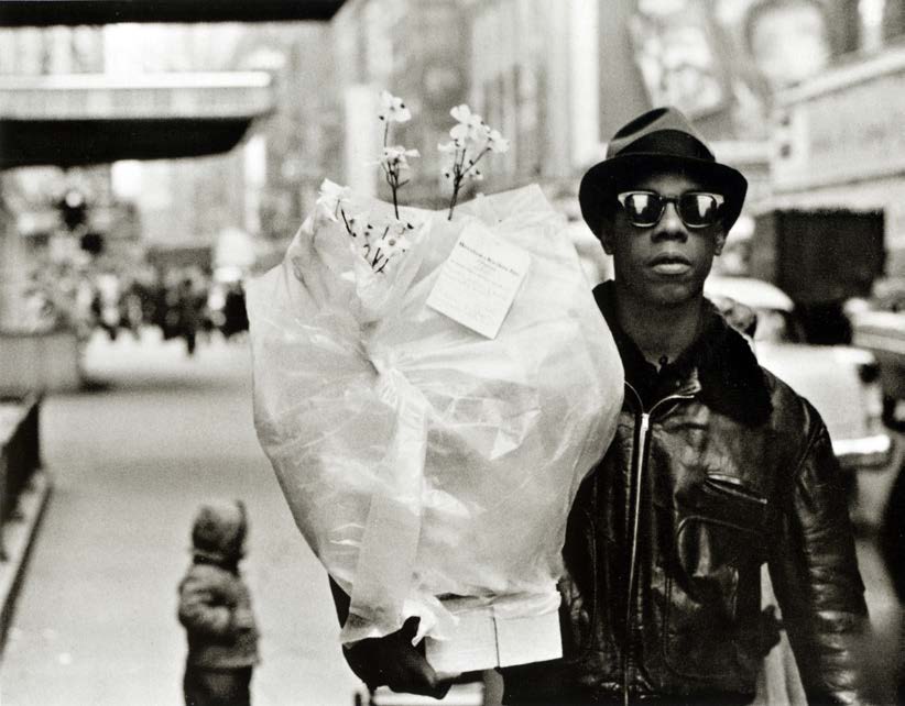 A Black man in street fashion holds a bouquet of flowers wrapped in plastic.