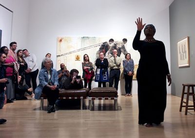 A group of people in a gallery room watch a woman perform.