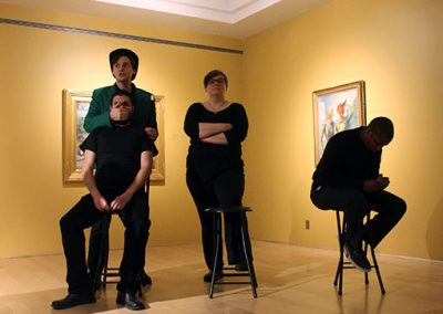 Two people in all black sit on stools. One stool is empty. Two people stand behind the others; one covers the mouth of a person on a stool.