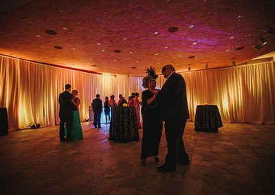 Couples in masquerade costumes dance. The ceiling has purple lights projected onto it.