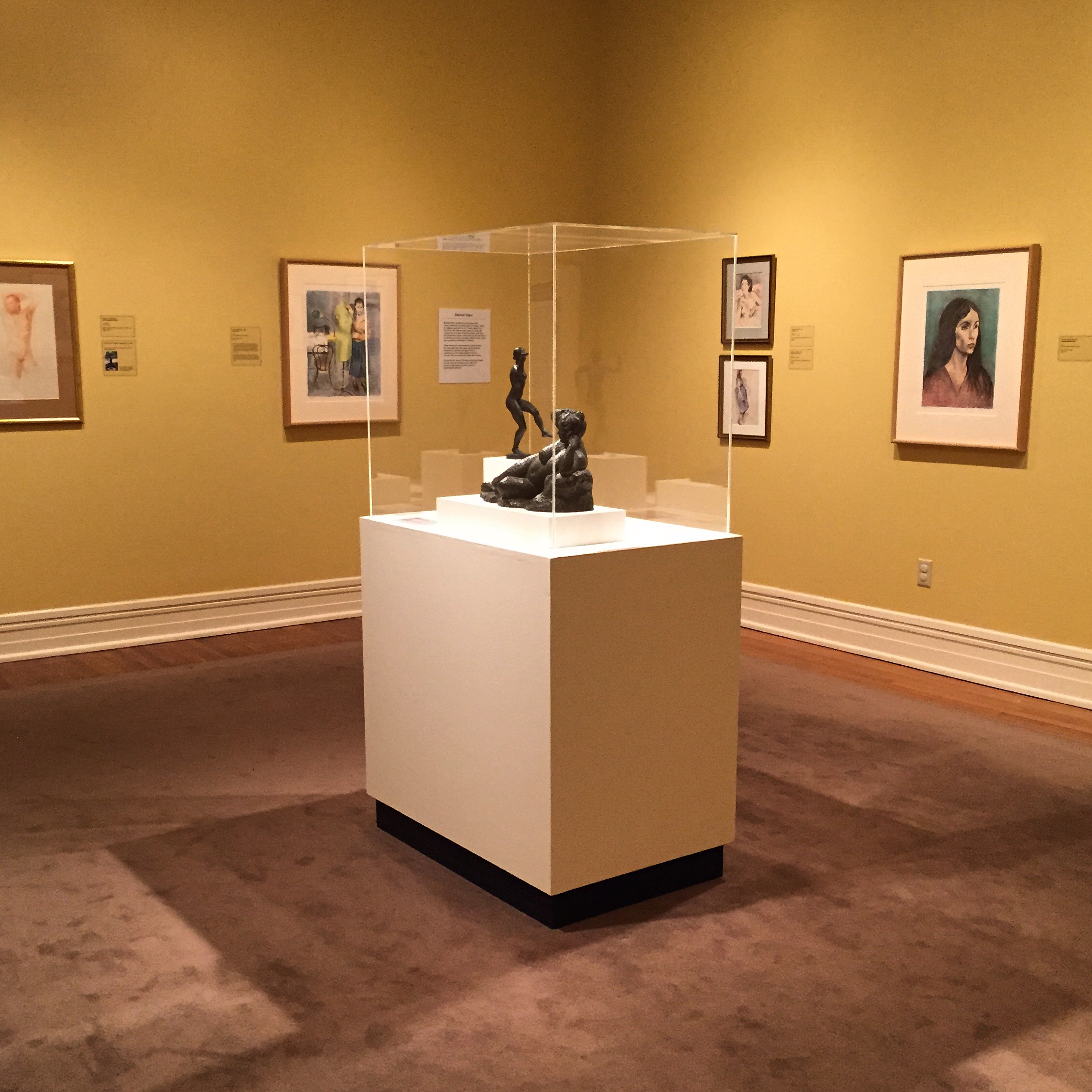 A gallery room with paintings on the wall and a glass case with a small sculpture.