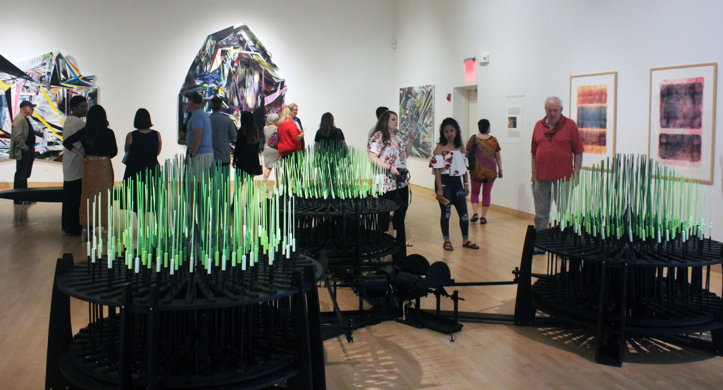 People view a gallery with 3D pieces that are large and circular with protruding slender blue and green rods.