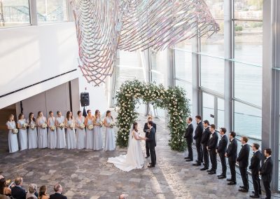 A wedding ceremony in the Hunter Museum lobby. The guests are seated, the bride and groom are at the altar with their bridesmaids and groomsmen. The image is taken from above so a large hanging sculpture is in view.