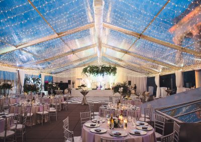 Wedding reception setup with an overhang of sheer fabric and warm string lights. Tables are in view. but the camera is pointed at the dance floor and band setup.