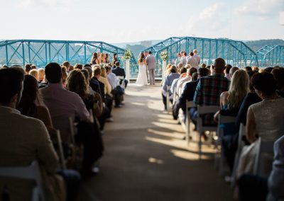 A bride and groom at the altar on the balcony of the Hunter Museum. The image is taken from the back of the aisle and includes the seated guests, bridal party, and the pedestrian bridge.