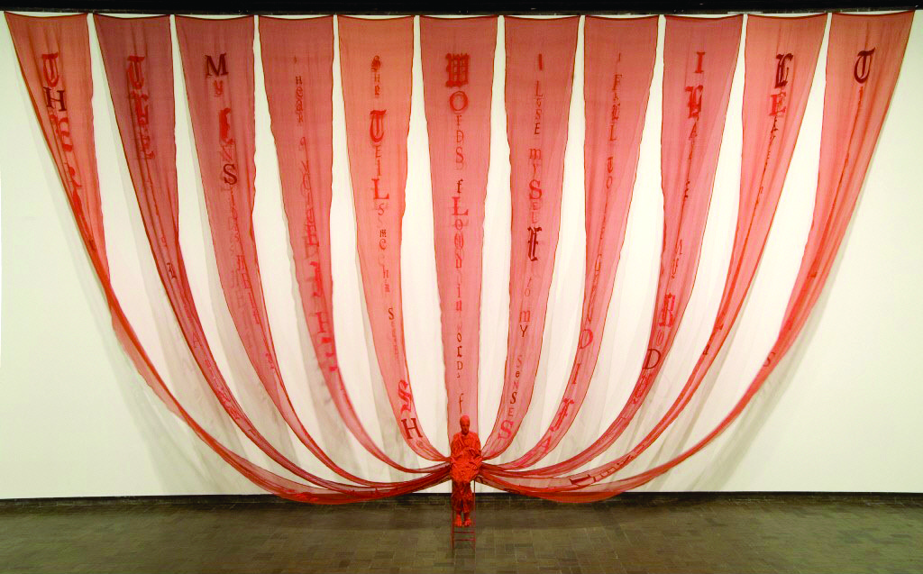 Leslie Dill's installation. A figure wrapped in red cloth sits as multiple red trains become red fabric banners connected to the top of the wall.