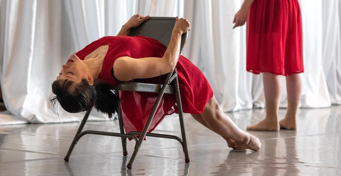 Two dancers in red dresses. One dancer is laying on her back stretched on a chair with her feet pointed.